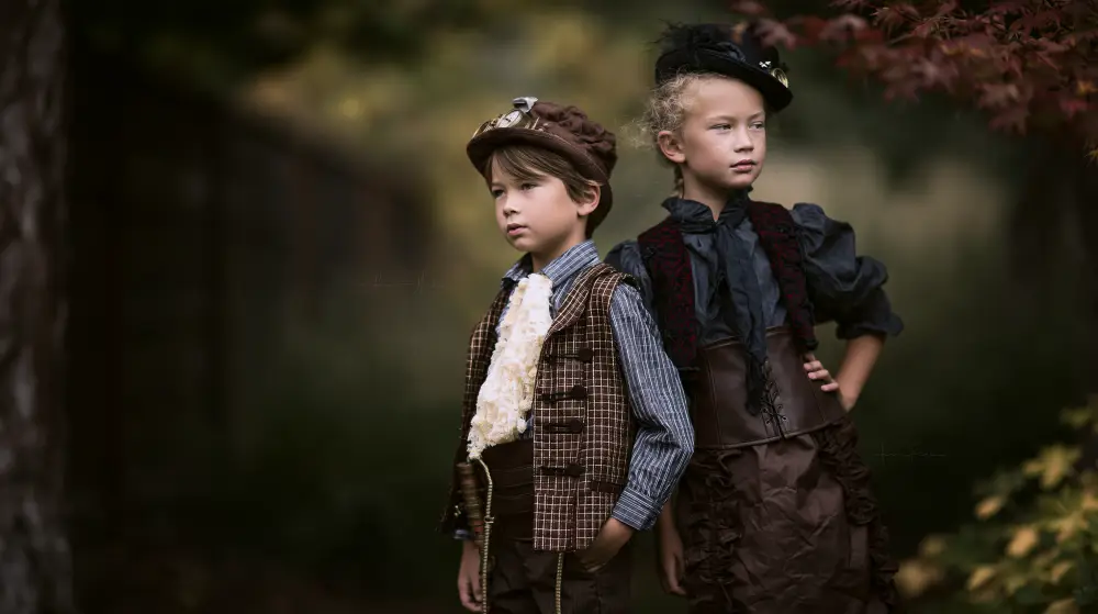 Two children wearing elegant steampunk Victorian clothing pose together in a quiet autumn garden.