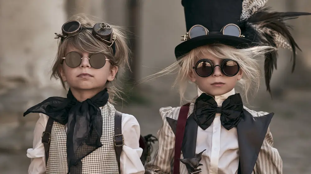 Two children wearing Victorian steampunk clothing with round goggles and a tall top hat pose for a portrait.