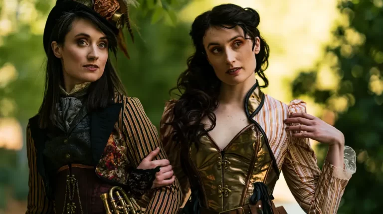 Two women wearing elegant steampunk Victorian clothing with polished brass corsets pose together in a forest setting.